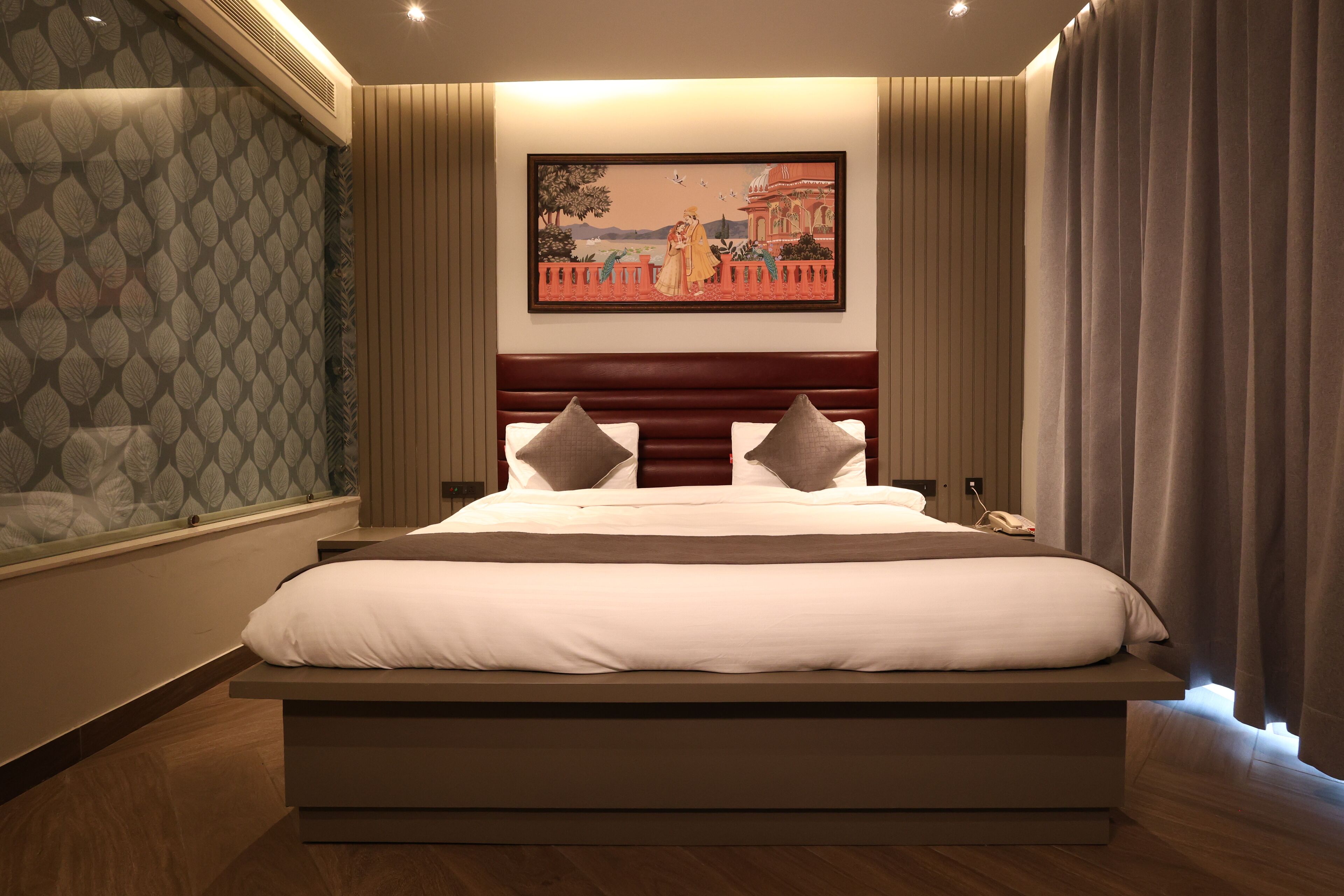 A modern hotel room with a large bed, a headboard with a decorative panel, a wall-mounted painting, and a window with a patterned curtain.