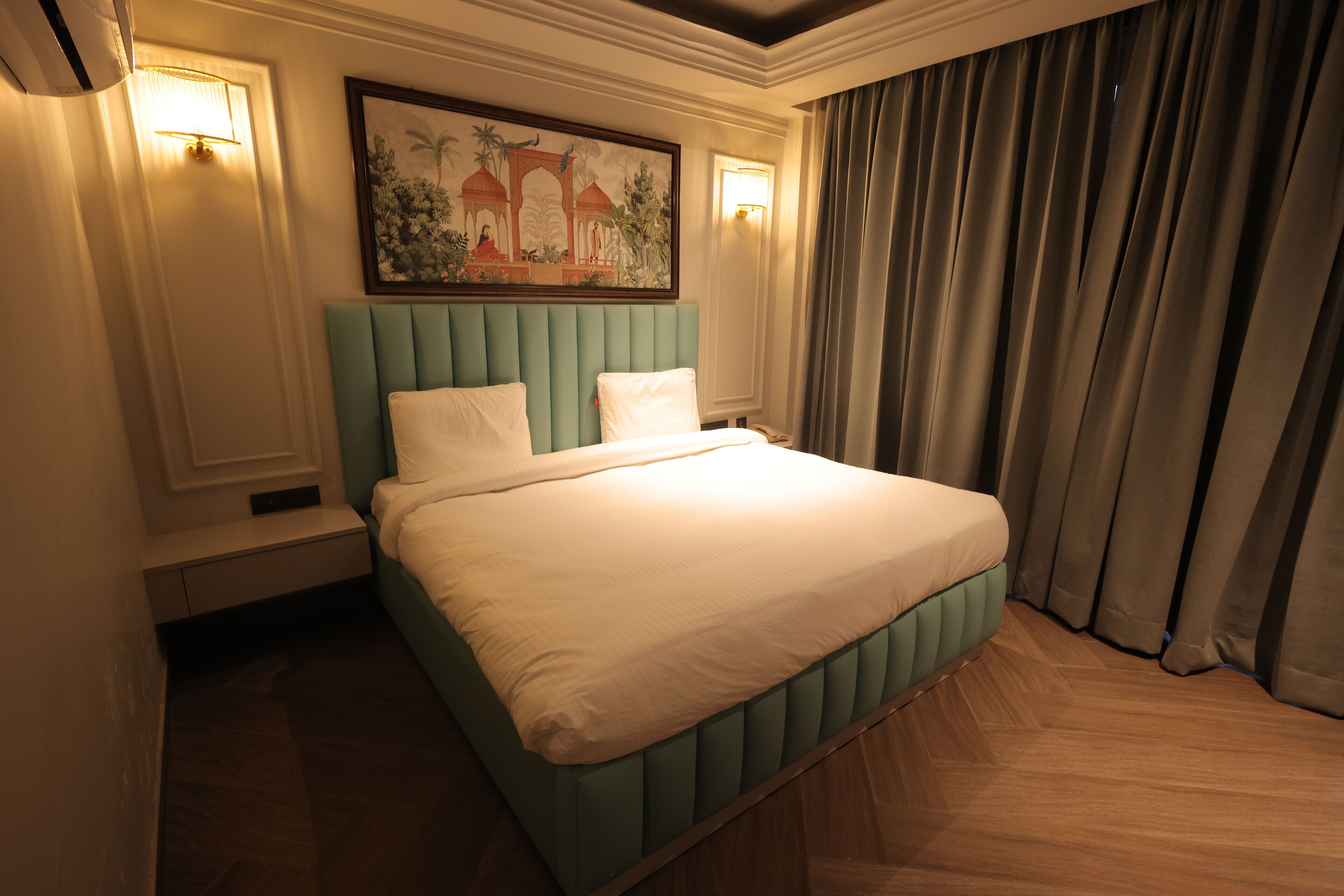 A hotel room with a large bed, a headboard with vertical panels, a wall-mounted painting, and a window with curtains.