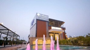 Un hotel moderno con un letrero llamativo, una piscina con fuentes de agua y una zona cubierta con estructura metálica.