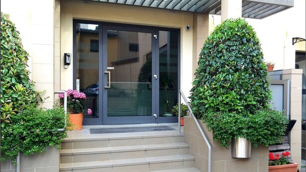 A building entrance with a glass door, steps leading up to it, and potted plants on either side. - 01. Apartment (Signa)