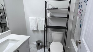 Ein kleines Badezimmer mit Toilette in Weiß, einem schwarzen Metallregal und einem weißen Waschbecken.