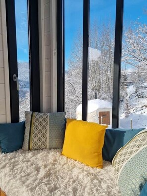Ein gemütlicher Sitzbereich mit einem flauschigen weißen Teppich, einer Couch mit gemusterten Kissen und Blick auf eine verschneite Landschaft durch große Fenster.