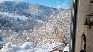 Una habitación con un amplio ventanal que da a un paisaje nevado con montañas y cielo despejado. En el interior, hay dos sillas de estructura metálica y una guitarra apoyada contra la pared.