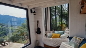 Una habitación acogedora con un ventanal grande que ofrece vistas panorámicas de un valle y montañas. Una guitarra negra cuelga de la pared, y un sofá mullido con cojines estampados se encuentra cerca del ventanal.