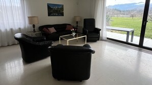 Un salon moderne avec un canapé, des fauteuils, une table basse et une vue sur un paysage verdoyant grâce à de grandes fenêtres.