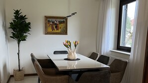 A dining area with a white table, six chairs, a vase with flowers, a painting on the wall, a potted plant, and a window with curtains.