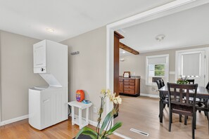 A room with a white washing machine, a small white table with a bottle, and a dining area with a wooden table and chairs. - Pastoral Tranquil Walkable to Trails Dining Shopping Secure Bike Boat Storage (Cape Elizabeth)