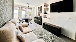A modern living room with a sectional sofa, a dining area with a view, a flat-screen TV, and a bookshelf.
