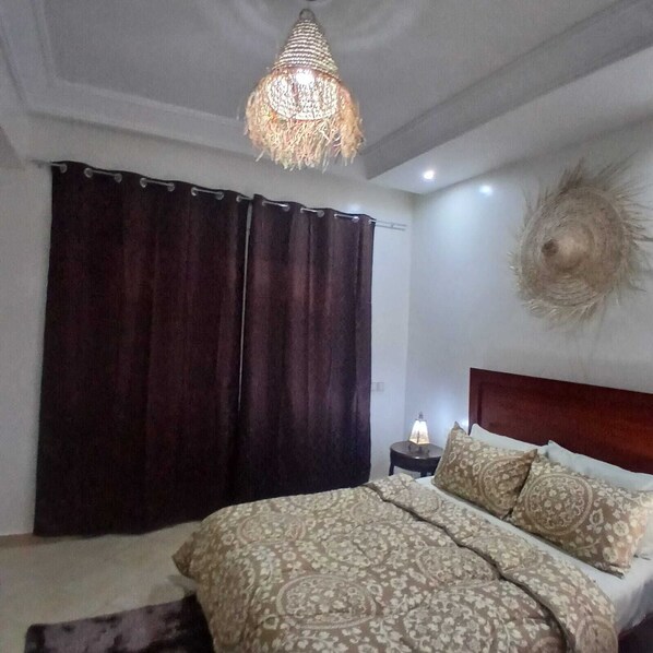 A bedroom with a bed, dark curtains, a patterned bedspread, a decorative wall hanging, and a hanging light fixture.