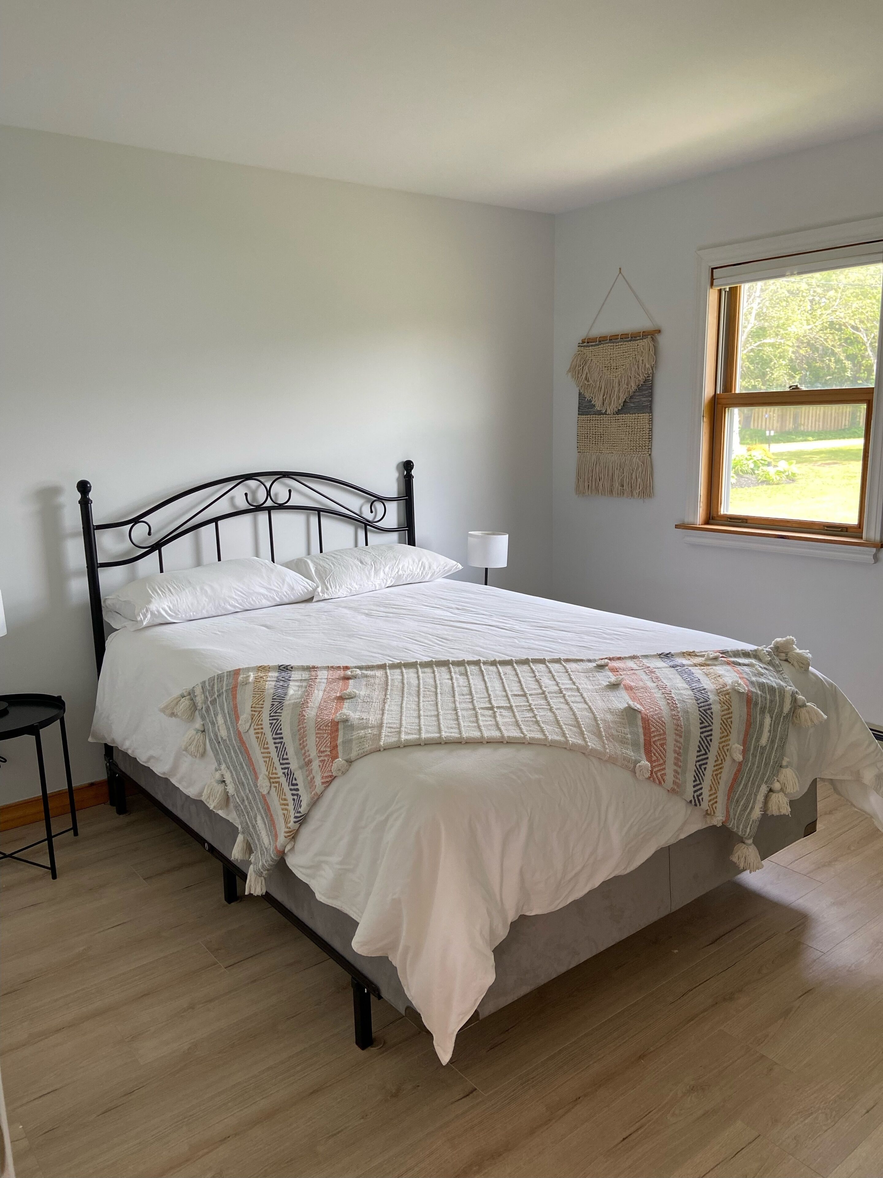 A bedroom with a bed, a bedside table, a hanging macrame wall decoration, and a window with a view of greenery.
