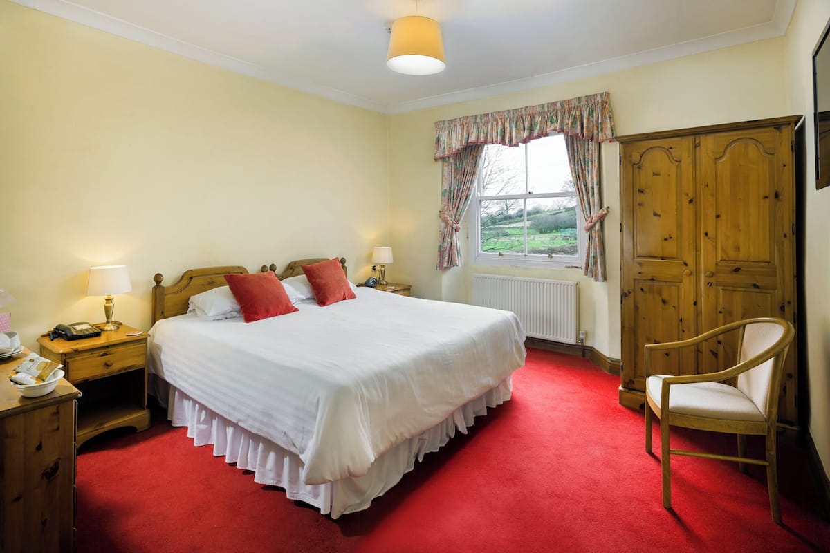 A bedroom with a large bed, a wooden wardrobe, a chair, a nightstand, and a window with curtains.