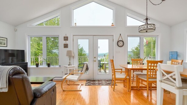 A spacious living room with a large TV, wooden flooring, a cozy seating area, and a dining set. - Oceanview Cottage on Cabot Trail - Main Level (Englishtown)