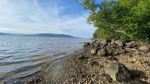 A rocky shoreline with clear water, a distant mountain range, and dense green foliage. - Oceanview Cottage on Cabot Trail - Main Level (Englishtown)