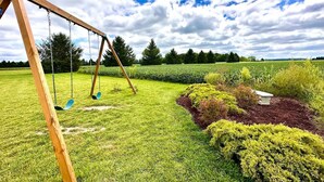 A wooden swing set in a grassy field with a hedge and a stone bench. - The Barn at Bloom & Bower (Risingsun)