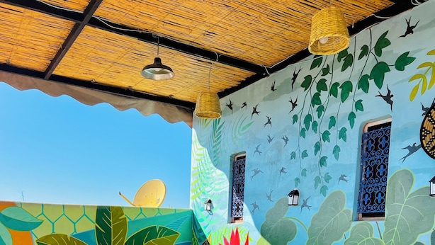 Une pièce lumineuse et accueillante, avec un canapé vert, des coussins aux couleurs vives et une fresque représentant des feuilles et des oiseaux.