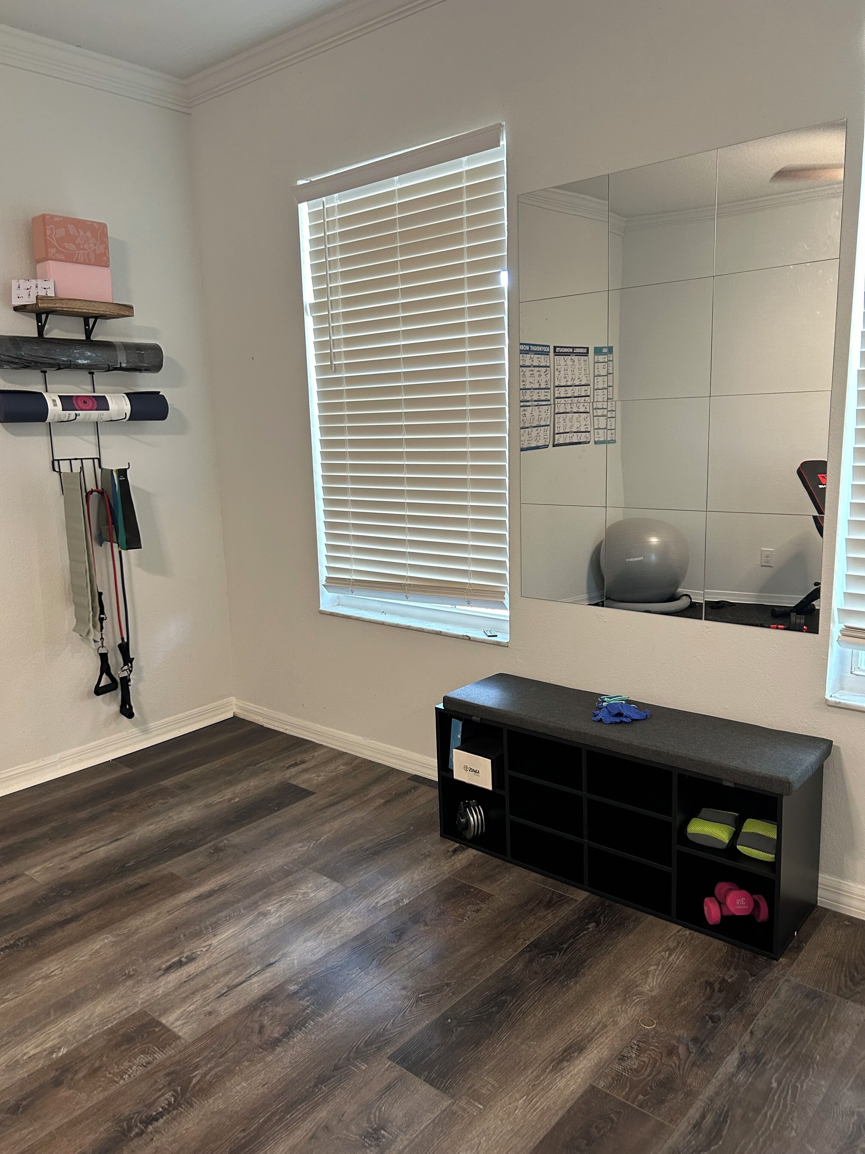 A room with a wooden floor, a black storage bench with labeled compartments, a large mirror, and a window with blinds.