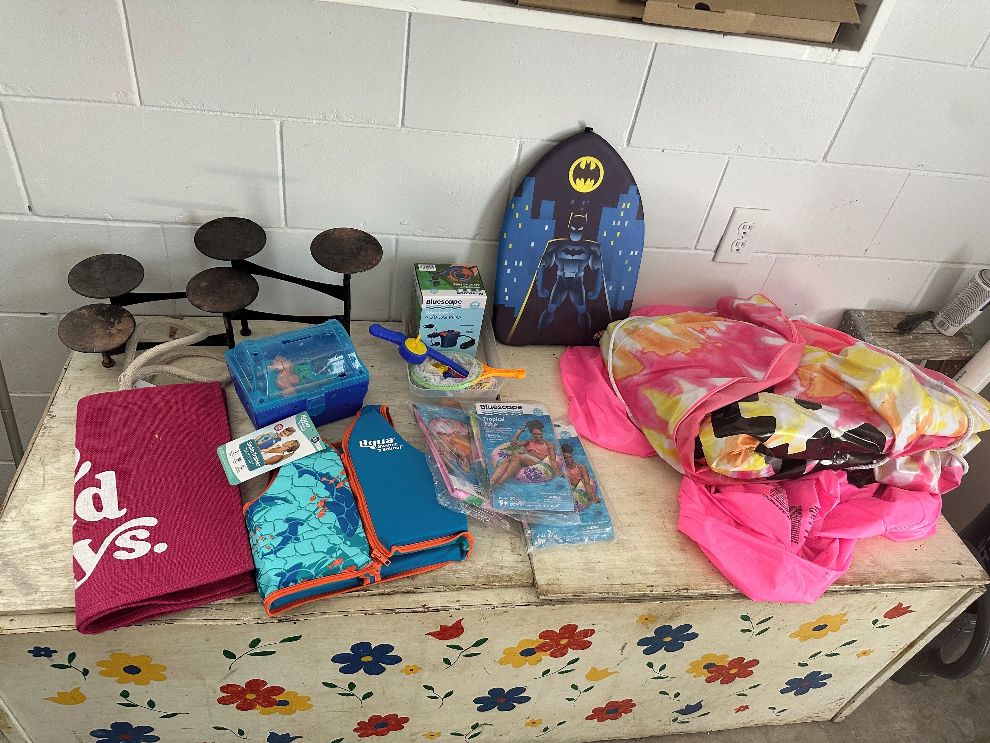 Une table sur laquelle se trouvent divers objets, notamment une boîte de rangement bleue, un sac rose, une planche de surf aux couleurs de Batman et une nappe à motifs floraux.