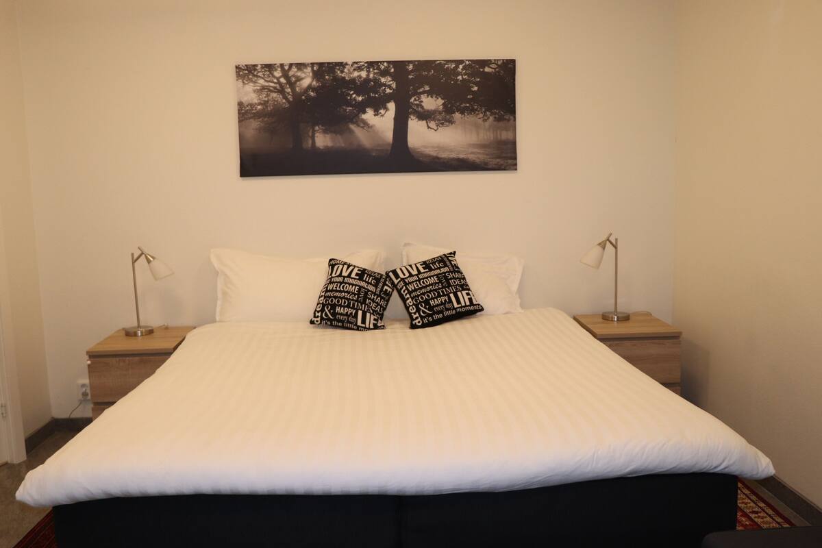 Una cama bien hecha con ropa de cama blanca y dos almohadas con texto motivacional, una mesita de noche con lámpara y un cuadro de un árbol colgado en la pared.
