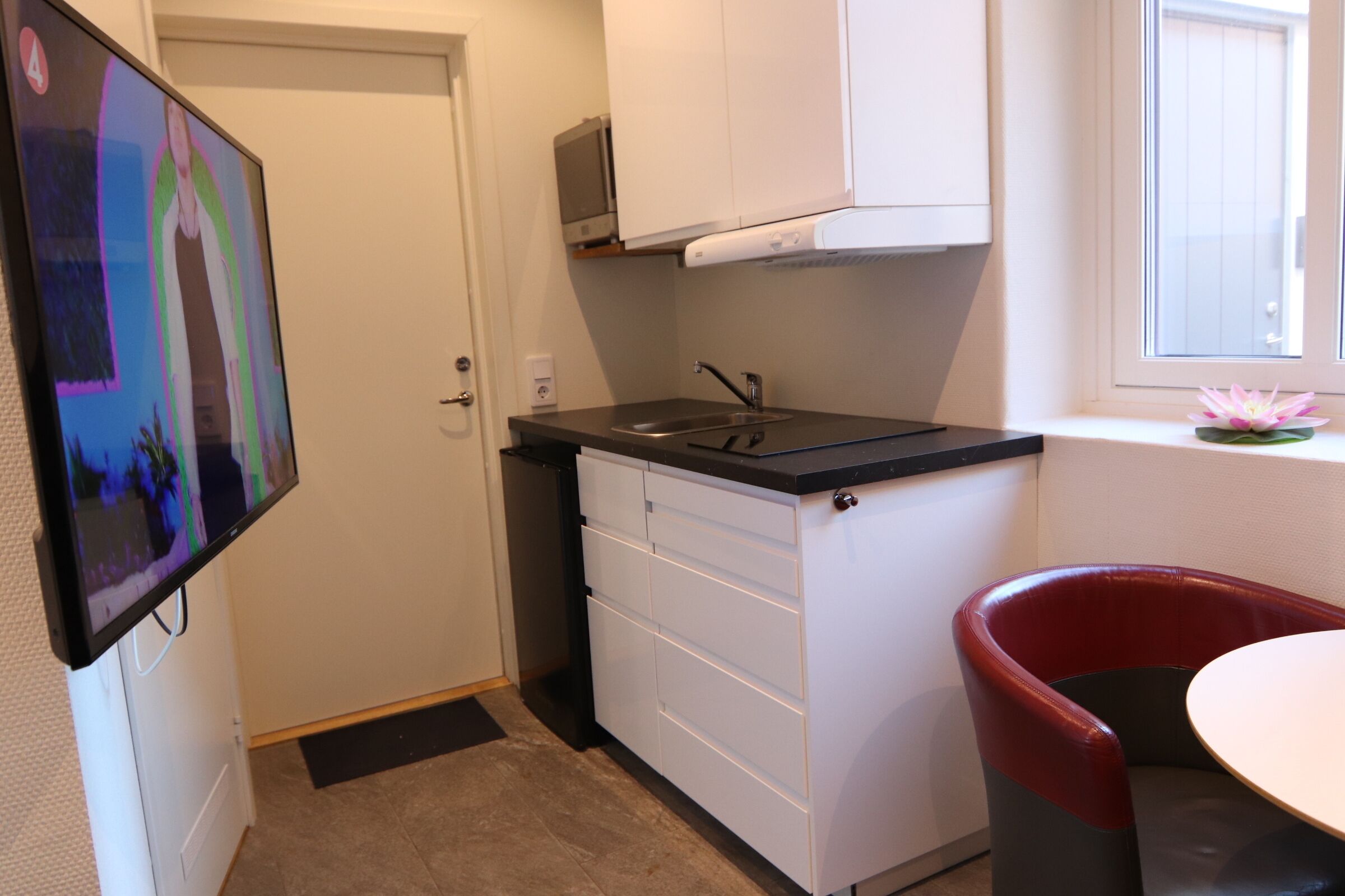 Cocina compacta con estufa integrada, armarios blancos, un lavamanos pequeño y una silla roja.