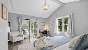 Een slaapkamer met een groot bed, een bureau en een stoel. Er is een raam met gordijnen, een plafondlamp en uitzicht op groen buiten.
