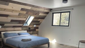 A bedroom with a sloped ceiling, a bed with blue bedding, a grey ottoman, and a window with a view of trees.