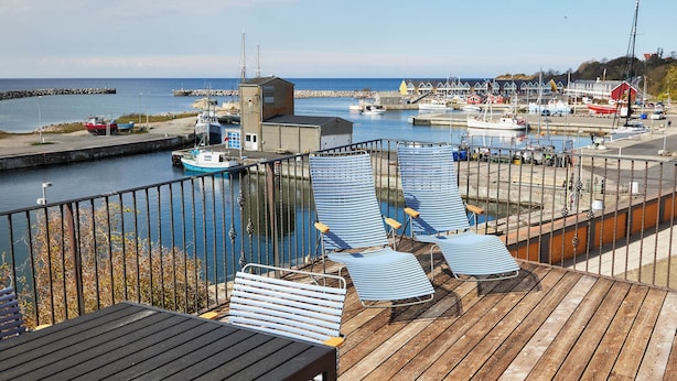 Une terrasse en bois avec des chaises longues et une table, donnant sur une marina où sont amarrés des bateaux et se trouve un quai.
