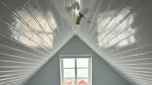 Une chambre avec un plafond en pente, une fenêtre, un radiateur, un lit avec des oreillers et un ventilateur de plafond.