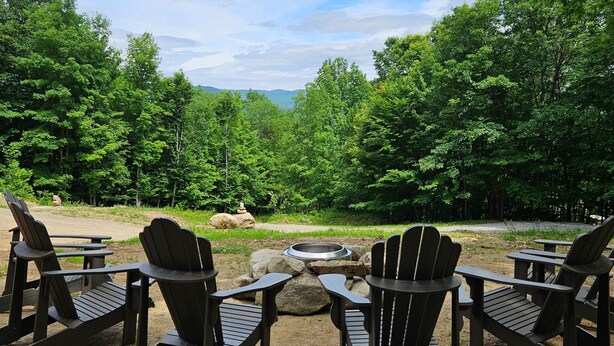 A campsite with Adirondack chairs arranged around a fire pit, surrounded by dense forest and mountains in the background. - Private, Mountain views, Close to Horse Show Grounds, Lake Placid and Hiking (Keene)