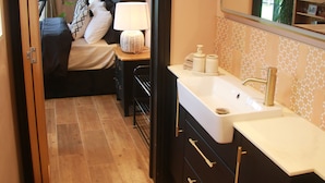 Habitación de hotel con suelo de madera, cama con ropa de cama blanco y negro, mesita de noche con lámpara, espejo grande y baño con lavabo y tocador.