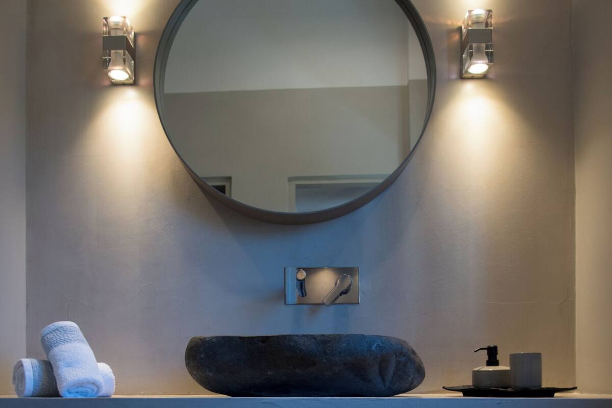 Un bagno con uno specchio tondo, due luci a parete, un portascopevoli con asciugamani, un dispenser per sapone e un lavandino con una vasca in pietra.