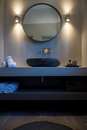A bathroom with a round mirror, two wall-mounted lights, a towel rack with towels, a soap dispenser, and a sink with a stone basin. - ILION Air Host - Near Athens International Airport (Markopoulo Mesogaias)