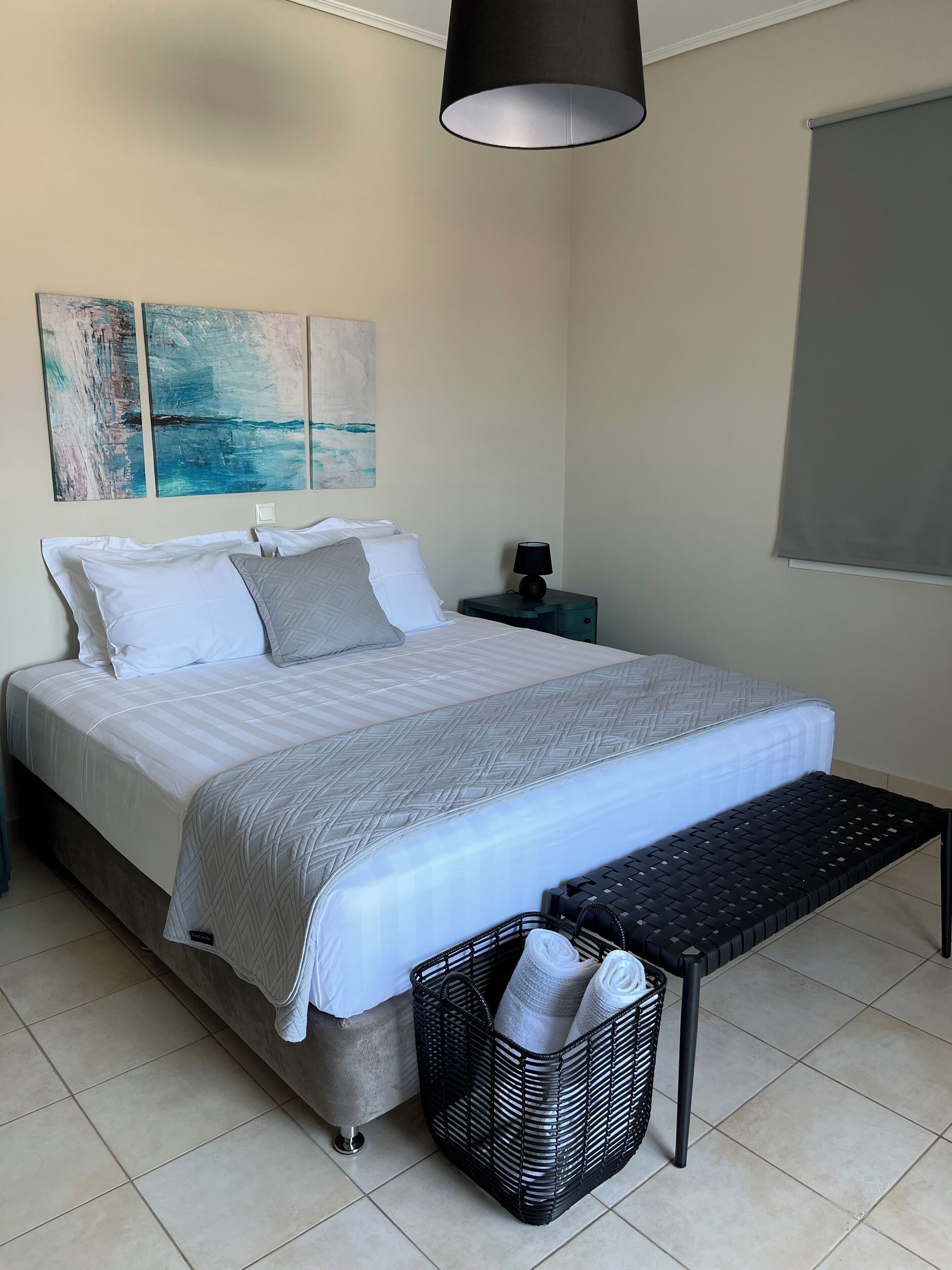 A neatly made bed with a grey and white duvet, a black bedside lamp, and a black woven bench.