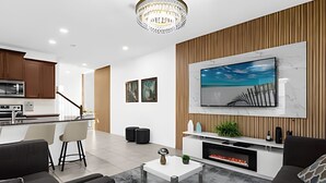A modern living room with a fireplace, a large TV mounted on the wall, and a kitchen in the background. - Amazing Home Solara Resort Comfort and Leisure (Kissimmee)