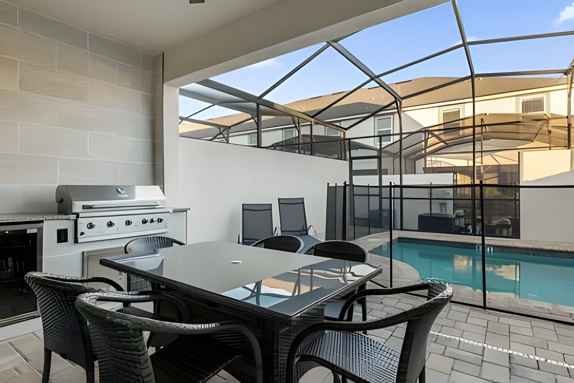 Een moderne buitenkeuken met een barbecue, eettafel en stoelen, met uitzicht op een zwembad en een woongebouw.