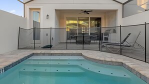 A swimming pool with a glass fence, a patio area with chairs and a table, and a building with a door and a ceiling fan. - Amazing Home Solara Resort Comfort and Leisure (Kissimmee)
