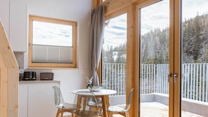 A cozy room with a wooden table and chairs, a small kitchen area, and a large window overlooking a snowy forest. - Rehfugium Lachtal by ALPS RESORTS (Oberwölz)