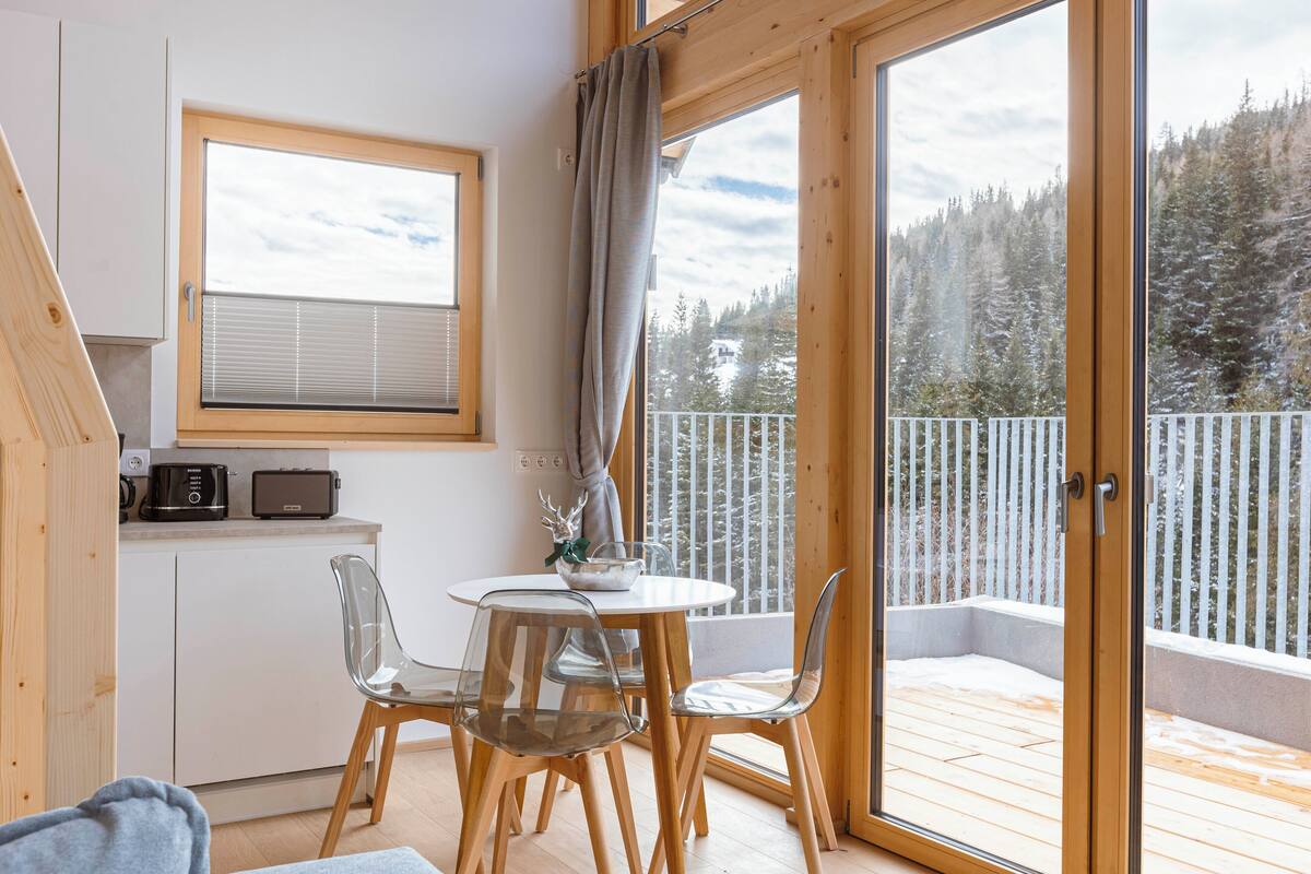Una habitación acogedora con una mesa y sillas de madera, un pequeño rincón de cocina y un amplio ventanal con vistas a un bosque nevado.