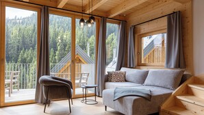 A cozy cabin with a sofa, a chair, and a small table. It has large windows with curtains, a balcony, and a view of a forest. - Rehfugium Lachtal by ALPS RESORTS (Oberwölz)