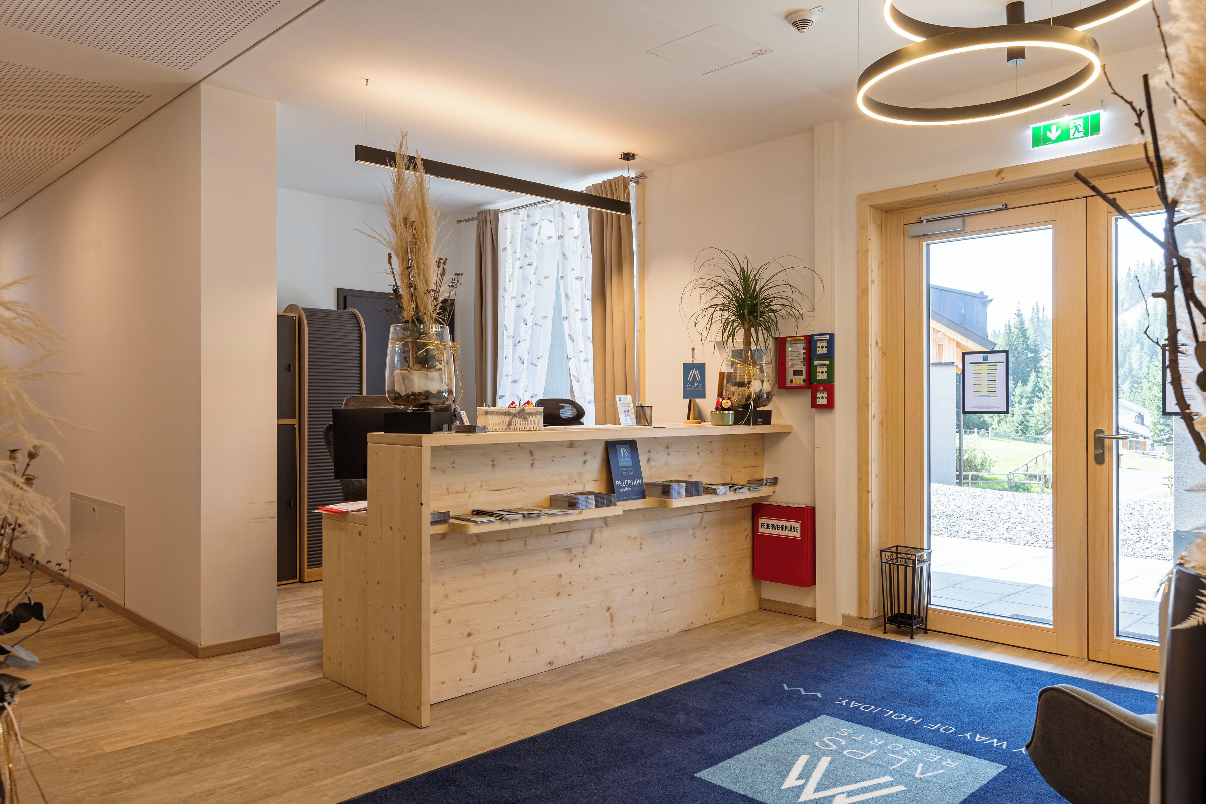 A reception desk with a wooden finish, a blue carpet with text, a potted plant, and a door leading outside.