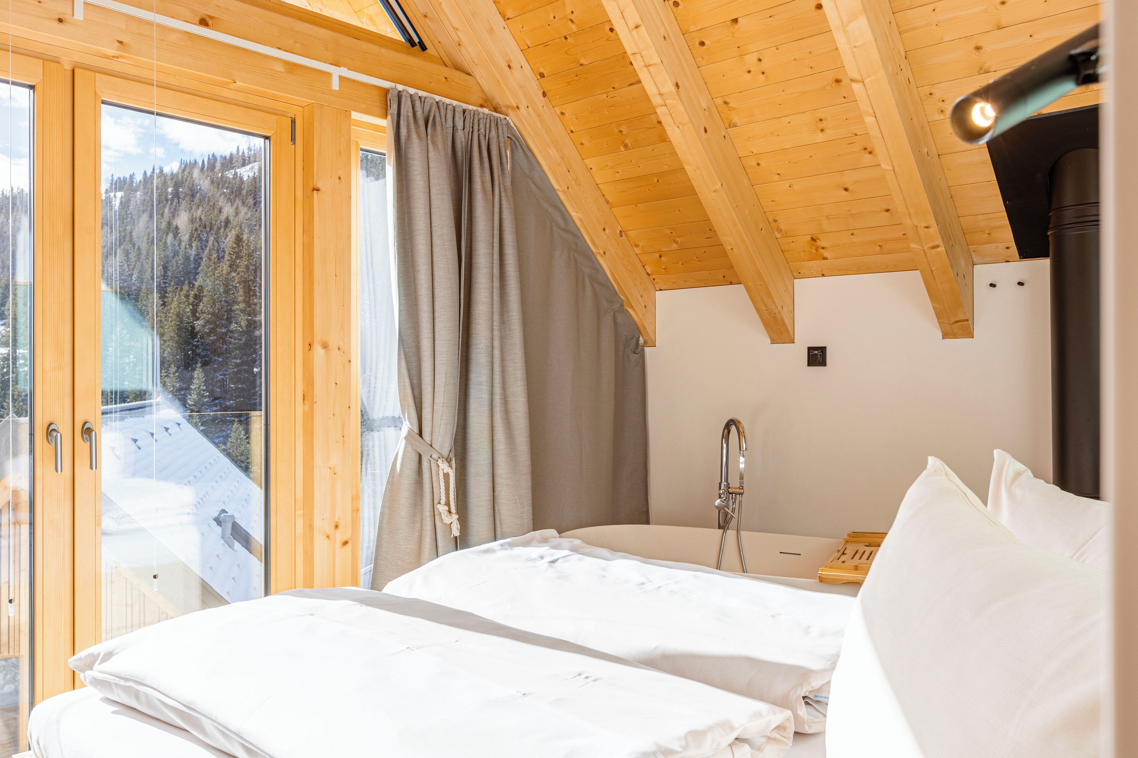 Ein Schlafzimmer mit Holzbalkendecke, einem Bett mit weißer Bettwäsche, einem Fenster mit Vorhängen und Blick auf eine verschneite Landschaft.