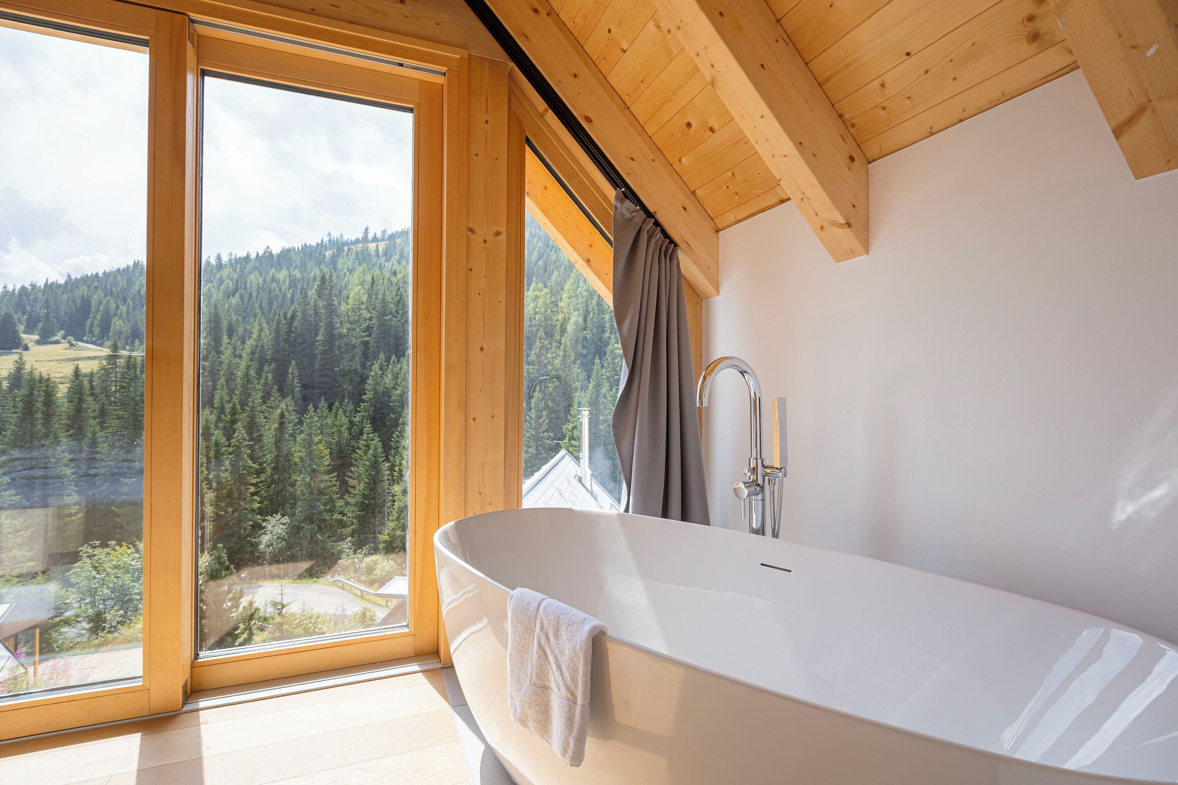 Un baño moderno con una bañera independiente, ventanas amplias con vista a un valle boscoso y vigas de madera en el techo.