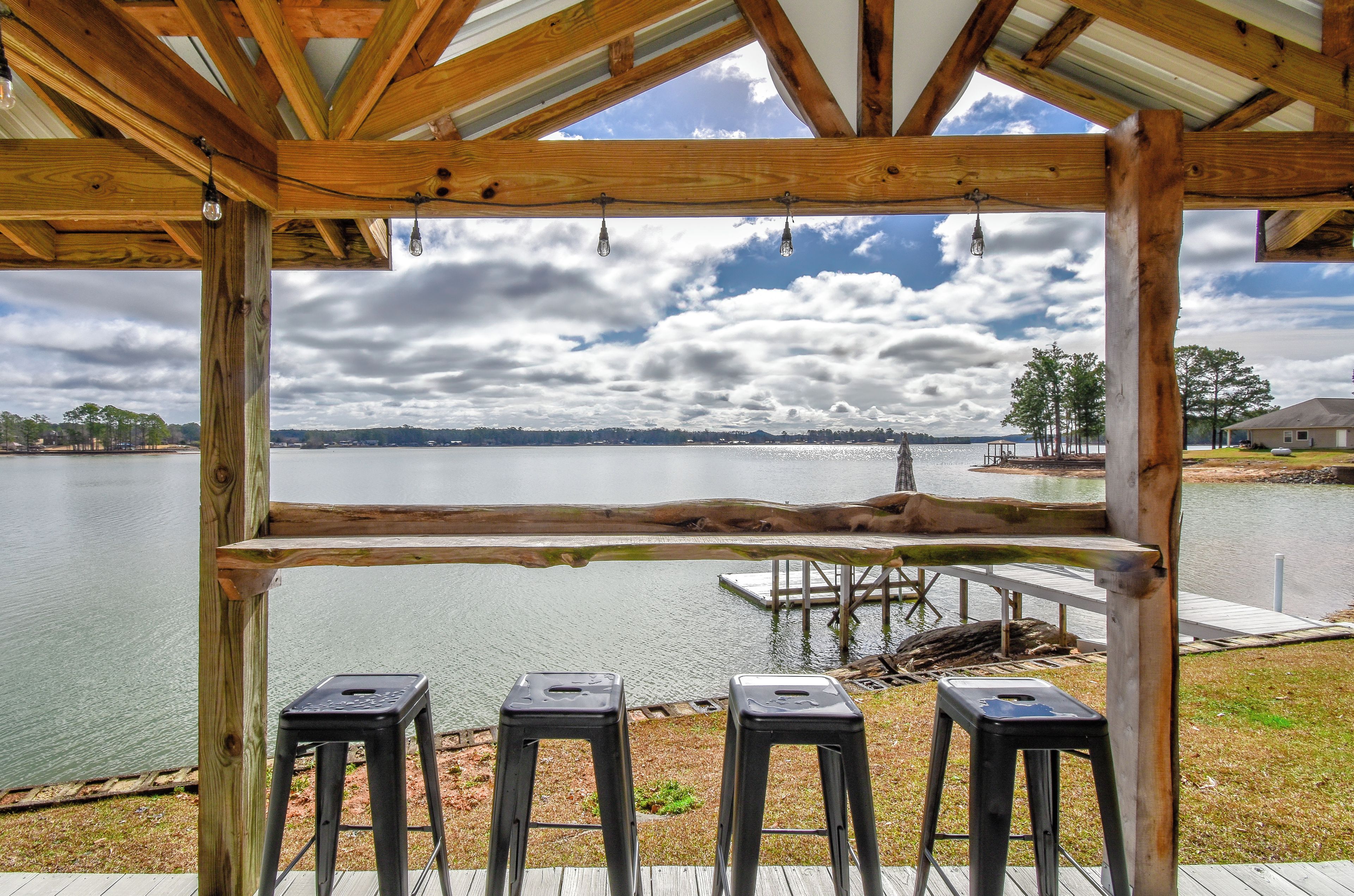 Une terrasse en bois offrant une vue sur un lac, trois tabourets et un quai.