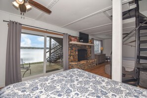Une chambre dotée d’un grand lit, d’une cheminée en pierre et offrant une vue sur un lac grâce à une porte-fenêtre coulissante.