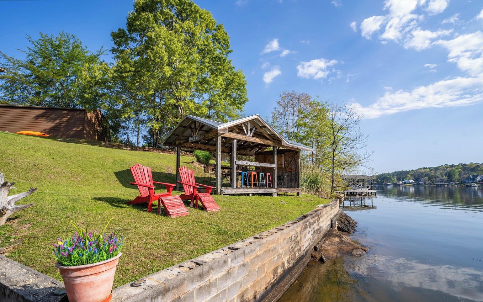 A lakeside cabin with a covered porch, red Adirondack chairs, and a potted plant.
