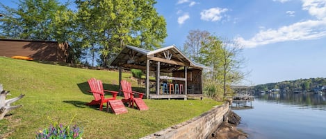 Una cabaña junto al lago con un porche cubierto, sillas Adirondack rojas y una planta en maceta.