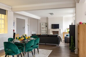 A modern living room with a dark grey sofa, a glass dining table with green chairs, and a flat-screen TV mounted on the wall. - 4-bdr Stunning Kensington Penthouse (London)