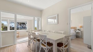 A spacious dining area with a large white table and white chairs, a view of the outdoors through large windows, and a modern interior design.
