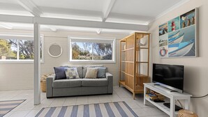 A living room with a grey sofa, a wooden bookshelf, a TV on a white stand, and a large window with a view of trees.