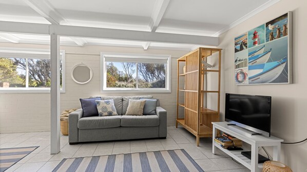 A living room with a grey sofa, a wooden bookshelf, a TV on a white stand, and a large window with a view of trees. - Beachhouse Currarong - Pet Friendly Beachfront (Currarong)