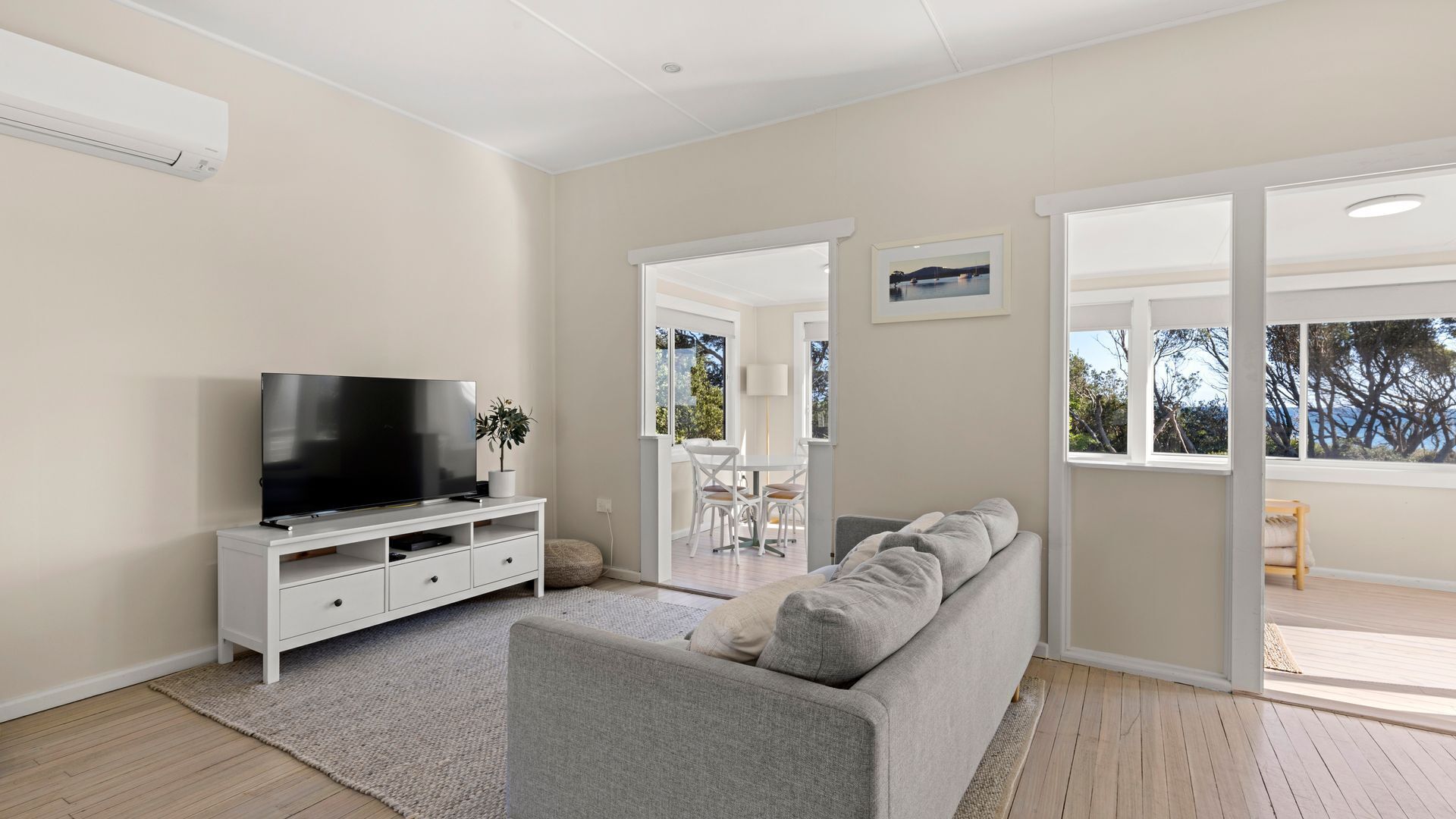 Un salon moderne avec un téléviseur à écran plat posé sur un meuble blanc, un canapé gris et un coin repas comprenant une table et des chaises.